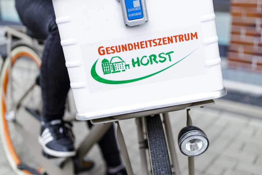 Person in rotem T-Shirt fährt mit einem weißen Botenfahrrad des Gesundheitszentrums Horst mit Kühlbox-Aufbau auf dem Gepäckträger.