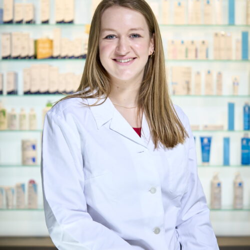 Porträt einer jungen Apothekerin im weißen Kittel vor einem Regal mit Kosmetik- und Apothekenprodukten